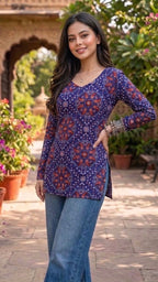 Woman wearing a patterned top and jeans standing in an outdoor setting with plants and flowers.