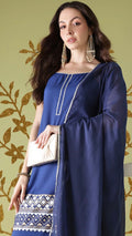 Woman wearing a blue traditional outfit with a white clutch against a decorative wall.