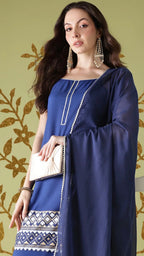 Woman wearing a blue traditional outfit with a white clutch against a decorative wall.