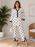 A woman models a matching two-piece lounge set featuring a white base with black circular patterns and black lace trim details, set against a neutral indoor background with a plant and wicker chair.